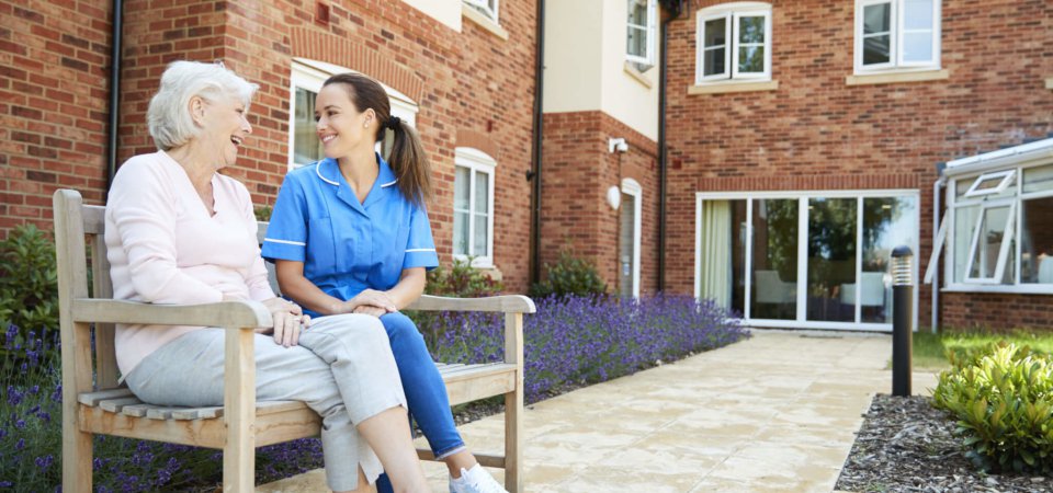 Senior Woman Sitting On Bench And Talking With Nurse In Retirement Home